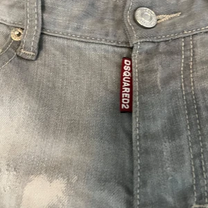 Grå slitna jeans från Dsquared2 - Säljer ett par grå jeans från Dsquared2 med slitna detaljer och färgstänk. Jeansen har raka ben, klassisk femficksmodell och knappgylf. Märkeslapp på baksidan och Dsquared2-tag vid fickan framtill.