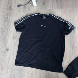 Svart t-shirt från Champion med logga - Svart t-shirt från Champion med vit logga på bröstet och längs axlarna. T-shirten har korta ärmar och detaljerade ränder samt Champion-tryck på ärmarna. Perfekt för en sportig och avslappnad stil.