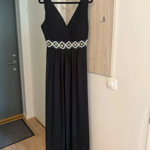 Balklänning - Elegant svart långklänning från Flower by Ohara med v-ringad omlott-topp och pärldetaljer i midjan. Klänningen har smala axelband och ett fint fall, perfekt för festliga tillfällen.