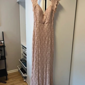 Ljusrosa långklänning med spetsdetaljer - Säljer en elegant ljusrosa långklänning med v-ringning och vackra spetsdetaljer. Klänningen har volang vid axlarna och ett feminint blommigt mönster i spetsen. Perfekt för speciella tillfällen.