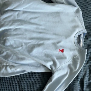 Ljusblå sweatshirt från Ami - Säljer en ljusblå sweatshirt från Ami med röd broderad logga (hjärta och A) på bröstet. Tröjan har rund halsringning och långa ärmar. Perfekt för en stilren och avslappnad look.