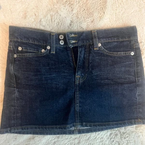 Mörkblå jeanskjol från SIS - Säljer en mörkblå jeanskjol från SIS, lågmidjad med dubbla knappar framtill och snygga kontrastsömmar. Kjolen är kort och har bakfickor med dekorativ söm. 