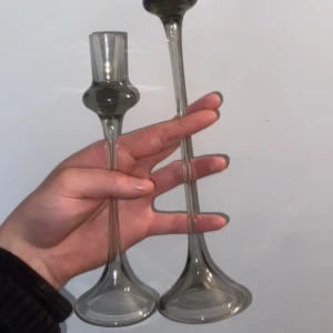 ELSA SÖDERBERG.2 ljusstakar - Bilden visar två eleganta ljusstakar i glas, hållna i en hand. Ljusstakarna har en modern design med smala, höga stjälkar och breda baser. Perfekta som inredningsdetalj för att skapa en mysig och stilren atmosfär hemma. Vill bara tillägga att båda två ljusstakarna ingår på köpet 