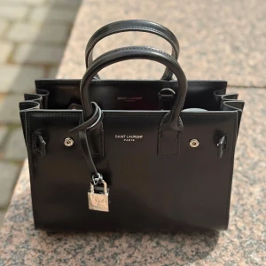 YSL Svart handväska från Saint Laurent - Perfekt storlek för dig som gillar att ha en mängd saker men i mindre väskor. Får plats med väldigt mycket. Plasten på låset är fortfarande kvar. Kommer med lås, nyckel, dustbag, kortet man får vid köp och förlängsningband som är helt oanvänd då jag själv aldrig använt det bandet. Väskan är i perfekt kondition då jag tar hand om mina varor med god hänsyn.                                                              Känn dig fri att skicka ett meddelande vid övriga frågor eller bilder! 