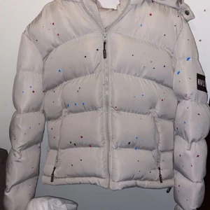 Vit pufferjacka med färgstänk från NVLTY - Säljer en vit pufferjacka från NVLTY med coola färgstänk i blått, rött och svart. Jackan har huva, dragkedja och en oversized look. Perfekt för att sticka ut i vinterkylan!