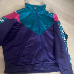 Retro träningsjacka från FILA - Säljer en färgstark retro träningsjacka från FILA i blått, turkost och rosa. Jackan har dragkedja framtill, hög krage och logotape längs ärmarna. Perfekt för dig som gillar vintage sportstil. Den är lite nopprig annars bra skick! Passar 36/38