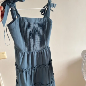 Blå klänning med knytaxelband från Pomelo - Säljer en ljusblå klänning från Pomelo i storlek XXS. Klänningen har knytbara axelband, smockad överdel och volangdetaljer. Perfekt för varma dagar.