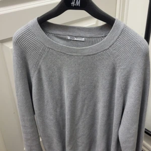 Ljusgrå stickad tröja från Zara - Säljer en stilren ljusgrå stickad tröja från Zara. Tröjan har rund halsringning och långa ärmar med ribbade detaljer vid axlar och ärmslut. Perfekt för lager-på-lager och enkel att matcha med olika outfits.
