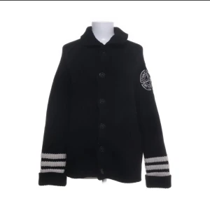 Svart stickad kofta med knappar från Stone Island - Svart stickad kofta med knäppning framtill och Stone Island-logga på ärmen. Koftan har ribbade muddar med två grå ränder på varje ärm och en klassisk krage. Perfekt för lager-på-lager och en stilren look.