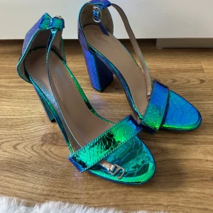 Blågröna klackade sandaler med ormskinnsmönster - Säljer ett par unika sandaler med hög klack i metallic blågrönt och ormskinnsmönster. Skorna har öppen tå, bred rem över foten och justerbart spänne vid ankeln. Perfekta för att sticka ut med en cool look. Oanvända storlek 36