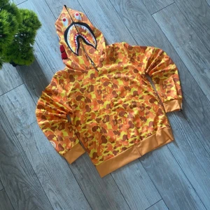 Orange camouflage hoodie med hajmotiv från BAPE - Säljer en orange camouflage hoodie från BAPE med hajmotiv på huvan och broderade bokstäver. Tröjan har dragkedja och långa ärmar, samt ribbade muddar och nederkant. Perfekt för dig som vill sticka ut med streetwear-stil.