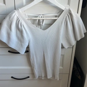 Vit ribbad topp med puffärmar från ONLY - Säljer en vit ribbad topp från ONLY med v-ringning och korta puffärmar. Toppen har knytning i ryggen och är tillverkad i ett stretchigt material som sitter snyggt på kroppen. Perfekt för dig som gillar en feminin och trendig stil.