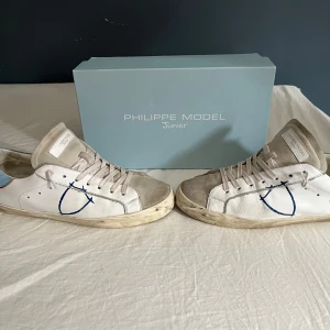 sneakers från Philippe Model  - Intressekoll på ett par snygga vita sneakers från Philippe Model Junior med beige mockadetaljer och blå broderad logga på sidan. Skicka gärna bud.