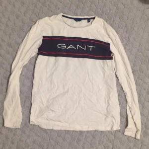 Vit långärmad tröja från GANT - Vit långärmad tröja från GANT med blått och rött band över bröstet där GANT-loggan är tryckt. Rund halsringning och klassisk passform. Perfekt för en stilren och avslappnad look.