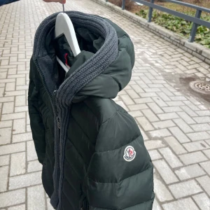 Mörkgrön dunjacka med huva från Moncler - Säljer en mörkgrön dunjacka från Moncler med stickad grå kant runt huvan och dragkedja framtill. Jackan har en klassisk pufferstil och Moncler-logga på ärmen. Perfekt för för alla årstider