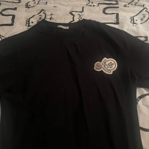 Svart t-shirt från Moncler med broderad logga - Säljer en svart t-shirt från Moncler med rund hals och en snygg broderad Moncler-logga på bröstet. T-shirten har en klassisk passform och är perfekt för dig som gillar stilrena plagg med exklusiv känsla.