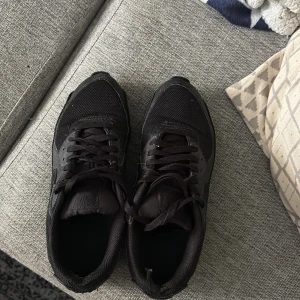 Svarta air Max 90 - Säljer ett par helsvarta Nike sneakers med klassisk design och snörning. Skorna har mesh- och syntetdetaljer för bra andningsförmåga och komfort. Perfekta för vardagsbruk eller träning. Snygg och stilren look som passar till det mesta.