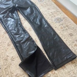 Svarta byxor i skinnimitation Nelly Trend  - Säljer ett par svarta byxor i skinnimitation med bootcut-passform. Byxorna har hög midja och dragkedja i sidan. Snygga och trendiga med en glansig finish som ger en cool look.