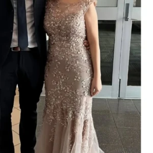 Champagnefärgad långklänning med spets och blommigt mönster - Elegant långklänning i champagnefärg med vackra spetsdetaljer och blommigt mönster. Klänningen har en figurnära passform, är ärmlös och har en rund halsringning. Perfekt för bal eller festliga tillfällen.