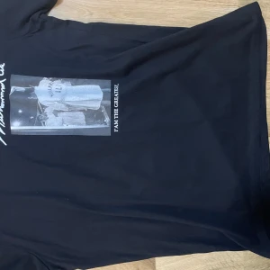 Svart Muhammad Ali t-shirt med tryck - Svart t-shirt med tryck av Muhammad Ali på både fram- och baksidan. På framsidan står det 'I AM THE GREATEST' och på baksidan finns ett större tryck med texten 'Muhammad Ali' och ett citat. Klassisk passform och rund hals.