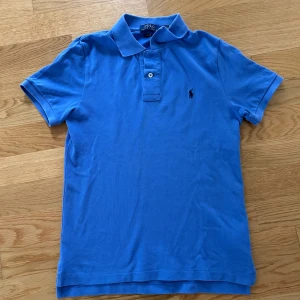 Blå pikétröja från Polo Ralph Lauren - Säljer en klassisk blå pikétröja från Polo Ralph Lauren i slim fit-modell. Tröjan är i bra skick och den är i storlek 14-16 vilket motsvarar XS.