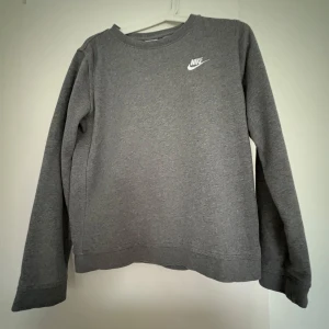 Grå sweatshirt från Nike - Säljer en klassisk grå sweatshirt från Nike med rund halsringning och vit Nike-logga på bröstet. Tröjan har långa ärmar och är tillverkad i mjukt bomullsmaterial. Perfekt för en avslappnad och sportig stil.