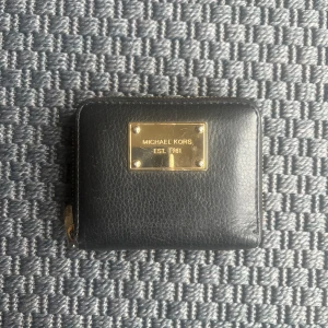 Svart plånbok från Michael Kors - Snygg svart plånbok från Michael Kors i skinn med guldfärgad metallplatta framtill. Plånboken har dragkedja runtom och flera fack för kort och mynt. Perfekt storlek för att ha med sig överallt.
