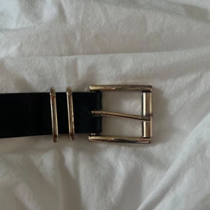 Svart bälte med guldfärgat spänne - Säljer ett stilrent svart bälte med guldfärgat metallspänne. Bältet har en klassisk design med två guldfärgade hållare och är gjort i ett slätt material med beige insida. Perfekt accessoar för att lyfta din outfit. Bältet är i acceptabelt skick men därför det billiga priset. Guld spännet har rostat lite och därav tappat färg men det är inget som syns jättetydligt på framsidan utan det syns mer på baksidan.😊