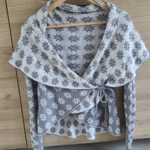 Gråvit omlottkofta med snöflingemönster från KappAhl - Supermysig stickad omlottkofta från KappAhl i grått och vitt med snöflingemönster. Koftan har långa ärmar, knytband i midjan och en stor krage som ger en extra cozy känsla. Perfekt för kyliga dagar!