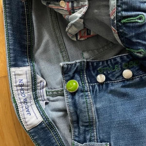 Blå jeans från Jacob Cohen - Säljer ett par klassiska blå jeans från Jacob Cohen med gröna detaljer och patch bak. Jeansen har raka ben och normal passform, med dragkedja och knapp framtill. Perfekta för dig som gillar stilrena och snygga jeans.