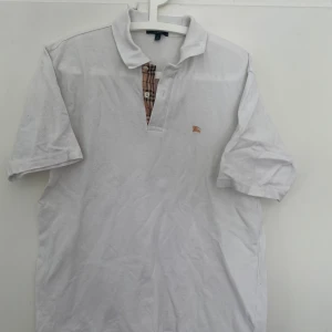 Vit pikétröja från Burberry - Vit pikétröja från Burberry med klassisk krage och knäppning med det ikoniska rutmönstret. Liten broderad logga på bröstet. Perfekt för en stilren och tidlös look. 