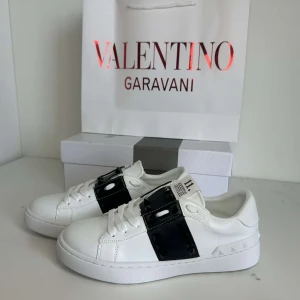 Valentino Garavani Skor - Säljer ett par vita sneakers från Valentino Garavani med bred svart rem över vristen och ikoniska nitar på hälen. Skorna har rund tå, platt sula och är tillverkade i skinn. Perfekta för dig som gillar stilrena men ändå unika detaljer.