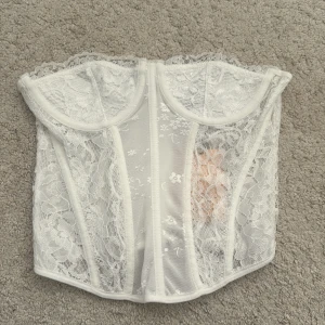 Vit spetskorsett med dragkedja - Säljer en vit korsett med vacker spets och blommigt mönster. Korsetten har bygel och dragkedja bak för enkel påklädning. Perfekt för att lyfta en outfit med en romantisk touch.