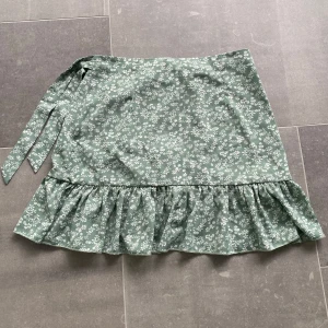 Grön blommig kjol med volang - Supersöt grön kjol med vitt blommönster och volang längst ner. Kjolen har omlottmodell med knytband i midjan och är perfekt för en somrig look.