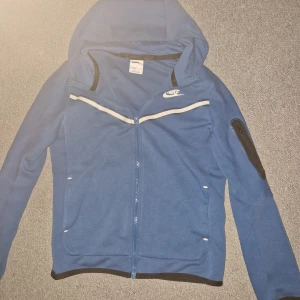 Blå hoodiejacka från Nike - Blå hoodiejacka från Nike med vit dragkedja och vita detaljer över bröstet. Jackan har huva, lång ärm och en ficka med dragkedja på ena ärmen. Perfekt för en sportig stil.