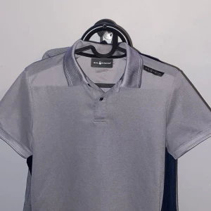 Ljusgrå pikétröja från Sail Racing - Säljer en ljusgrå pikétröja från Sail Racing i storlek XS. Tröjan har korta ärmar, klassisk krage och knappar vid halsen. Perfekt för en stilren och sportig look.