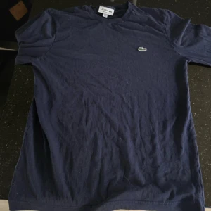 Mörkblå långärmad t-shirt från Lacoste - Säljer en mörkblå långärmad t-shirt från Lacoste med klassisk krokodillogga broderad på bröstet. Tröjan har en normal passform och är tillverkad i mjuk bomull. Perfekt för en stilren och avslappnad look.