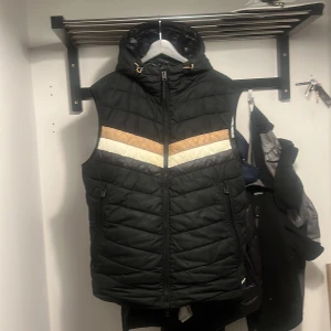 Svart dunväst från BOSS med huva - Snygg svart dunväst från BOSS med quiltad design och huva. Västen har dragkedja framtill, två sidofickor med dragkedja och coola detaljer i vitt och beige som går i ett V-mönster över bröstet. Perfekt för lager-på-lager och streetstyle. Köp för ca 1år sen. Original pris 3200kr