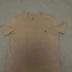 Beige t-shirt från Polo Ralph Lauren - Helt oanvänd Snygg beige t-shirt från Polo Ralph Lauren i mjuk bomull. Klassisk rund hals och korta ärmar. På bröstet finns det ikoniska broderade polospelaren i brunt. Perfekt basic med clean look och skön passform.