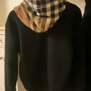 Svart Burberry hoodie med rutmönstrad huva - Svart hoodie från Burberry i storlek S med en ikonisk rutmönstrad huva i beige, svart och bruna toner. Tröjan har snörning vid halsen och långa ärmar. Perfekt för dig som vill ha en klassisk streetstyle med lyxig touch.