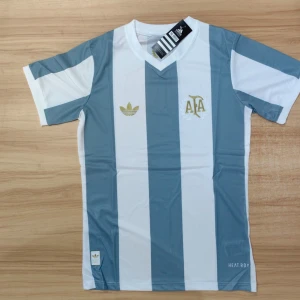 Argentina retro fotbollströja Adidas M - Snygg Argentina retro fotbollströja från Adidas i storlek M. Tröjan har breda vertikala ränder i ljusblått och vitt, guldiga detaljer och AFA-logga på bröstet. V-ringad hals och korta ärmar. Tillverkad i lätt, ventilerande polyester med HEAT.RDY-teknologi. Sprillans my kommer med förpackning