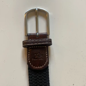 Svart flätat bälte från Billybelt - Snyggt svart flätat bälte från Billybelt med detaljer i mörkbrunt skinn och silverfärgat spänne. Klassisk design med diskret logga på lädret 👍 