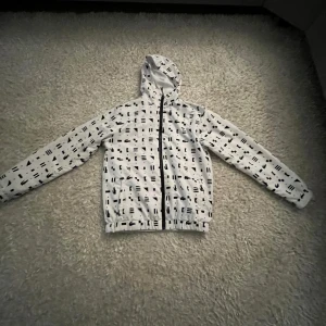 Vit vindjacka med svart mönster - Snygg vit vindjacka med svart abstrakt mönster och huva. Jackan har hel dragkedja framtill och resår vid ärmslut och midja. Perfekt för blåsiga dagar och enkel att matcha med streetwear eller casual outfits.