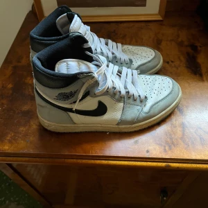 Nike Air Jordan 1 High vit/svart/grå - Nike Air Jordan 1 High i vitt, svart och ljusgrått skinn med klassisk svart swoosh och vingar-logga på sidan. Högt skaft, perforerad tå och vita snören ger en ikonisk streetstil. Perfekta för dig som vill sticka ut med en tidlös sneaker.