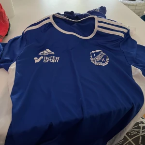 Blå Adidas fotbollströja IK Vändelsö - Snygg blå fotbollströja från Adidas med vita detaljer och tryck från IK Vändels samt Intersport. Tröjan har korta ärmar, rund hals och klassiska Adidas-ränder på axlarna. Perfekt för träning eller match. Materialet är lätt och andas bra.
