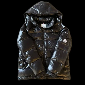 Svart Moncler dunjacka med huva - Snygg svart dunjacka från Moncler med glansig finish och klassisk logga på ärmen. Jackan har huva, dragkedja framtill och praktiska fickor. Perfekt för kalla dagar när du vill hålla stilen och värmen.