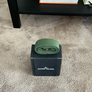 Grönt bälte från Stone Island - Snyggt militärgrönt bälte från Stone Island med matchande grön spänne och ikonisk logga på metallspännet. Köpt på NK i Stockholm är i strl. 03
