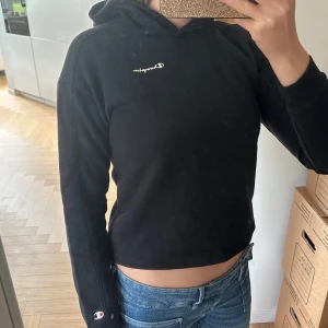 Svart hoodie från Champion - Svart hoodie från Champion med broderad logga på bröstet och ärmen. Klassisk modell med huva och långa ärmar, croppad passform som ger en modern vibe. Perfekt till jeans eller joggers för en chill look.