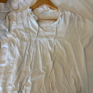 Vit blus med knyt och volang - Supersöt vit blus med korta ärmar, v-ringning och volangdetalj vid halsen. Blusen har knytband framtill och är gjord i ett lätt, krinklat bomullstyg som känns luftigt och somrigt. Perfekt för dig som gillar romantiska och avslappnade vibes.