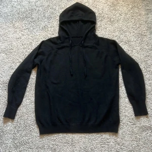 Svart hoodie med huva och snörning - Svart hoodie med klassisk huva och snörning framtill. Tröjan har långärmade ribbade muddar och är tillverkad i ett mjukt stickat material som känns skönt mot huden. Perfekt för chill dagar och enkel att matcha med jeans eller joggers.